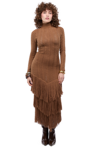Mujer usando un maxi vestido tejido en marrón con mangas largas, escote versátil y tiras amarrables. Su faldón tiene tres capas de flecos.