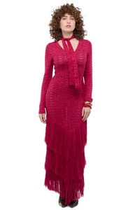 Mujer usando un maxi vestido tejido en rojo con mangas largas, escote versátil y tiras amarrables. Su faldón tiene tres capas de flecos.